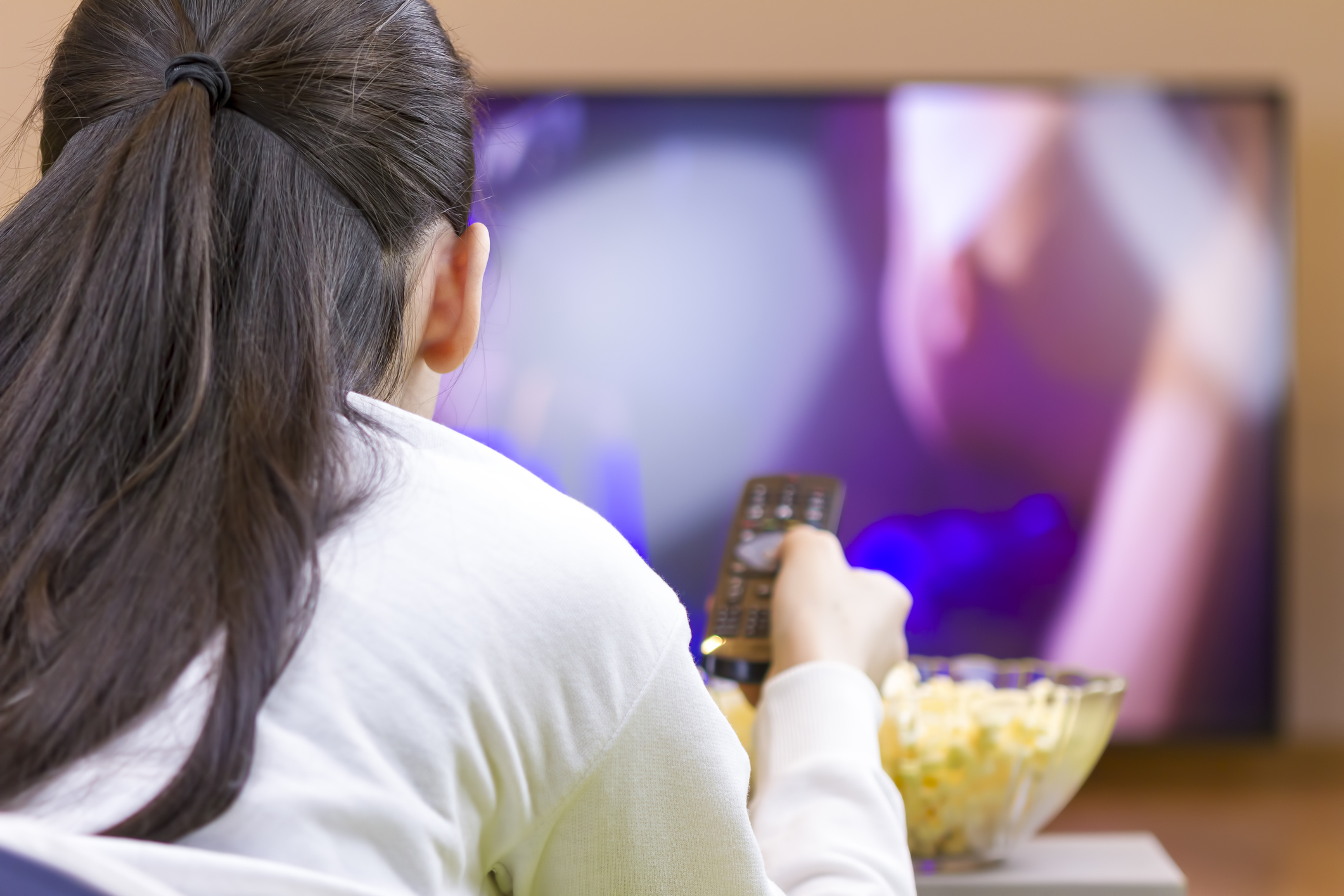 Teenager girl with remote control laying down and watching tv eating popcorn.
